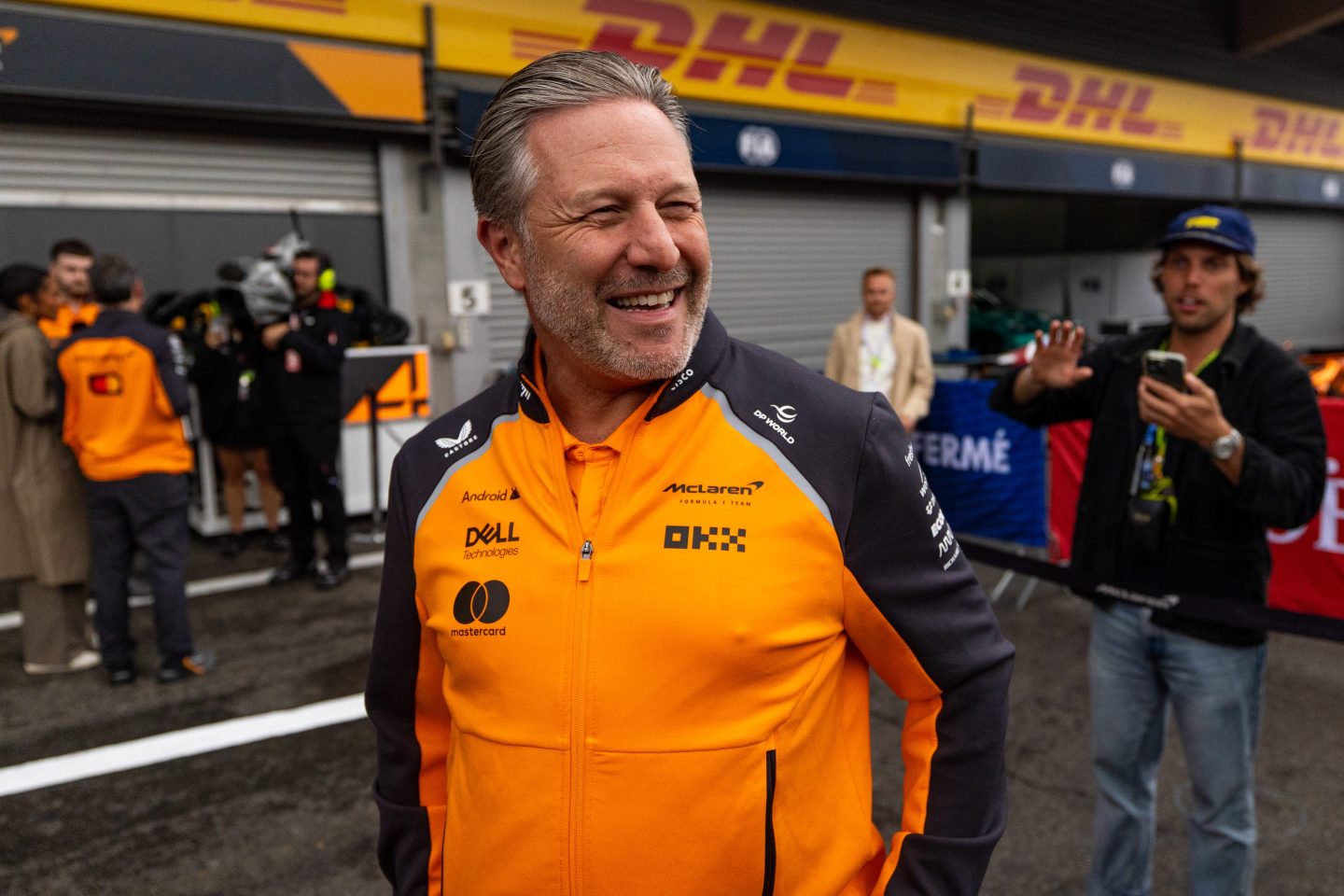 Zak Brown walks through the Formula 1 paddock, smiling.